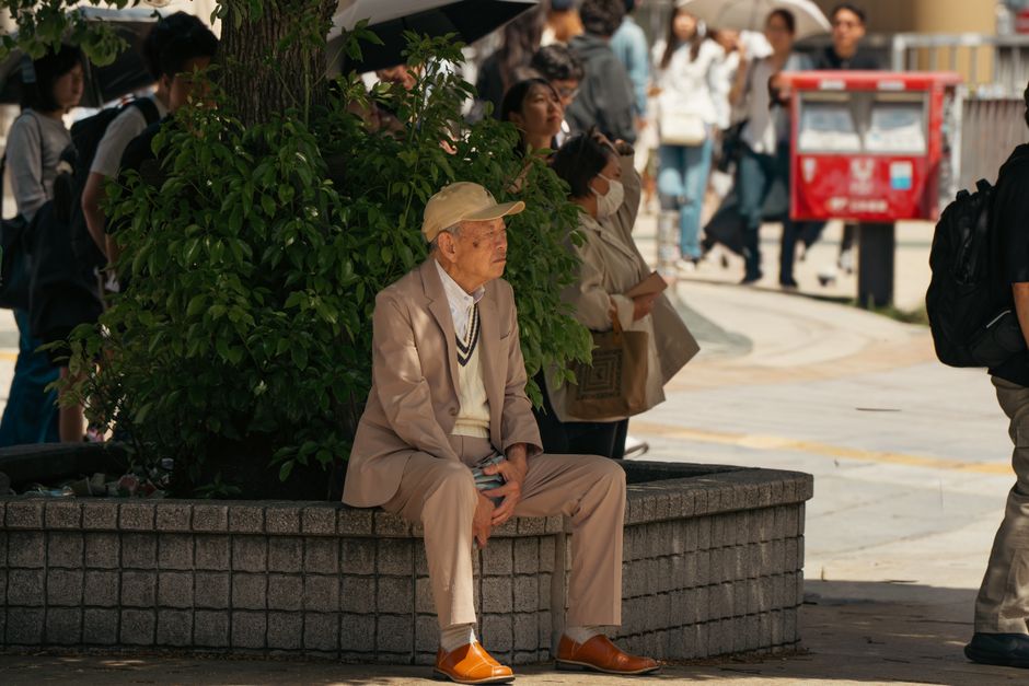 Elderly man sitting by plants in Tokyo streets, surrounded by pedestrians.