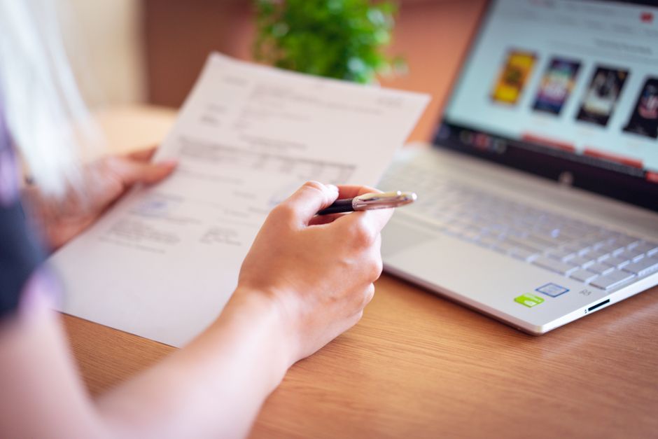 A person reviewing printed paperwork next to an open laptop at a kitchen table