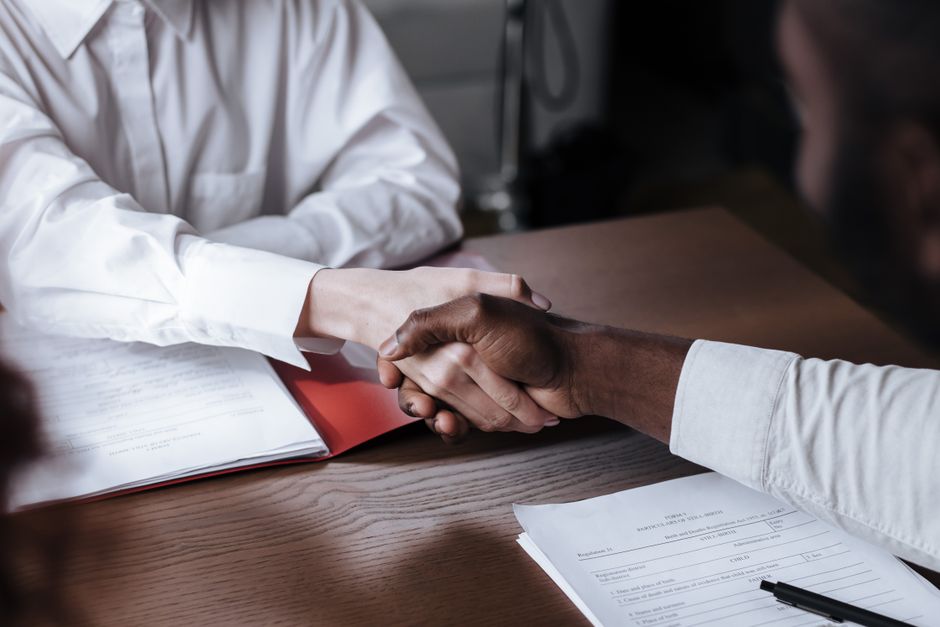 Two people shaking hands across a desk in an office interview setting